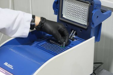 Siyah eldiven giyen bir laboratuar teknisyeni örnekleri mavi bir bilimsel alete yerleştiriyor, muhtemelen termal döngü ya da PCR makinesine.
