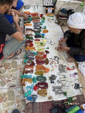 Satılık renkli boncuklar ve mücevherlerle çevrili yerde oturan iki kişinin yer aldığı bir piyasa sahnesinde çeşitli tasarım ve malzemeler sergileniyor.