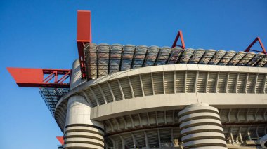 Benzersiz bir mimari tasarımı olan modern bir stadyum yapısı, açık mavi gökyüzüne karşı kıvrımlı bir cephe ve kırmızı aksanlar içeriyor.