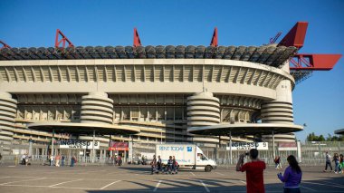 Modern mimarisi olan büyük bir spor stadyumu, açık mavi bir gökyüzü, önünde yürüyen insanlar ve yakınlarda park etmiş bir teslimat minibüsü.