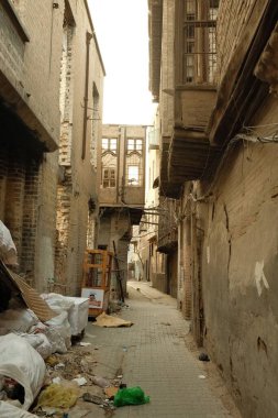 Eski binalar arasında dar bir ara sokak. Çürüme ve ihmal belirtileri var. Yere çöp ve enkaz saçılmış.