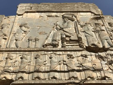 Gökyüzünün altında duran figürlerden haraç alan kralın oturduğu bir kraliyet sahnesini tasvir eden antik taş tasviri.