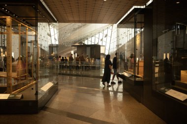 Müze içi cam vitrinler, ziyaretçiler, sıcak ışıklandırma ve yansıtıcı döşemeler; modern mimari tavan ve büyük pencereler