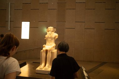Müzenin içinde oturan antik heykel ve onu izleyen iki ziyaretçi, sıcak aydınlatma