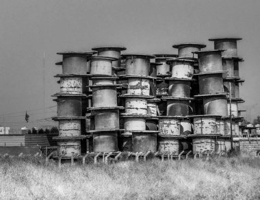 Endüstriyel bir alanda istiflenmiş silindirik beton makaralar, yıpranmış yüzeyler ve soyulmuş boya, siyah-beyaz atmosferik bileşim