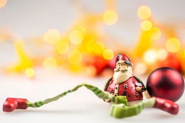 Una santa claus de cerámica y una bola roja de Navidad sobre fondo blanco con luces de Navidad amarillas borrosas — Foto de Stock