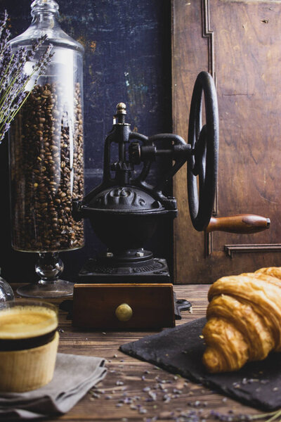 Старинная кофемолка на темном фоне с чашкой кофе и круассаном