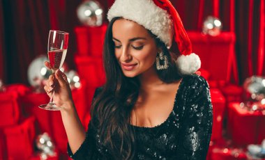 Mutlu yıllar sana! Noel Baba şapkalı, bir bardak şampanyayla güzel, seksi esmer kadın. Yeni yıl partisi. Noel arifesi. Kırmızı hediye kutuları ve disko toplarının arka planında.