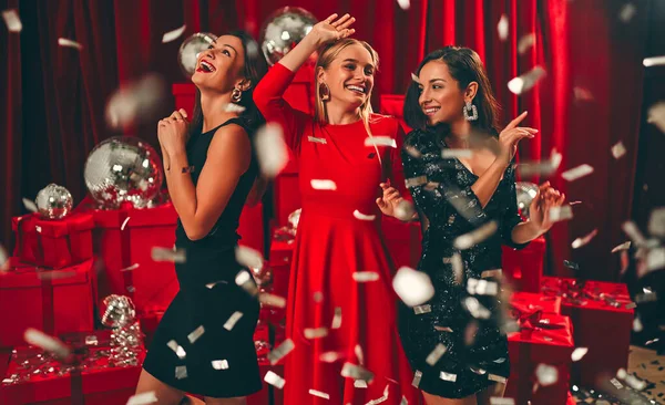 Happy New Year to you! Three beautiful sexy women dancing in confetti. New Year\'s party. Christmas Eve. Against the background of red gift boxes and disco balls.