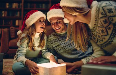 Mutlu Noeller ve mutlu yıllar, mutlu aile Noel Baba şapkalarıyla yeni yılı bekliyor. Aileler sevimli kızlarına hediye paketi sunuyorlar. İçeriden sihirli ışık.