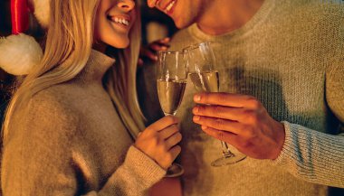 Cropped image of beautiful young couple in Santa hats is holding glasses of champagne and smiling while celebrating New Year at home.