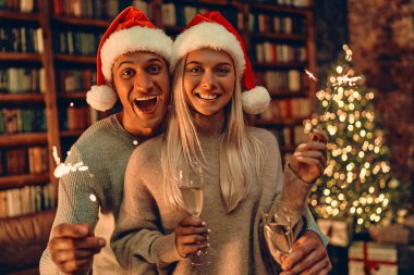 Beautiful young couple in Santa hats is holding glasses of champagne and smiling while celebrating New Year at home. Against the background of the Christmas tree.