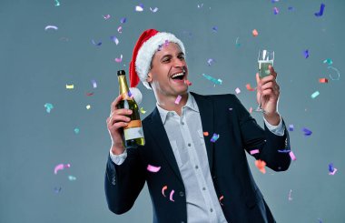 Successful man in a jacket with a bottle of champagne and a glass celebrates New Year. Studio photo on a gray background with confetti.