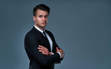 Serious businessman in a black suit isolated on gray background. Handsome man standing with crossed arms.