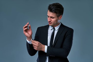 Portrait of trendy, attractive, stunning man in black tuxedo with tie fasten button on sleeve cuffs of his white shirt, isolated on grey background.