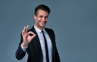 Everything is OK! Happy young man in black suit gesturing OK sign and smiling while standing against grey background.