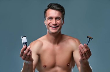 Handsome young man isolated. Portrait of shirtless muscular man is standing on grey background with trimmer in one hand and razor in other and smiling. Men care concept.