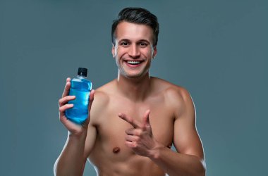 Handsome young man isolated. Portrait of shirtless muscular man is standing on grey background with bottle of mouthwash. Men care concept.