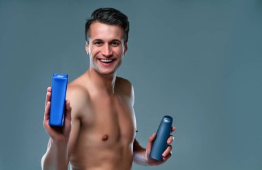 Yakışıklı genç adam izole edilmiş. Üstsüz kaslı adamın portresi gri arka planda elinde şampuanla duruyor. Erkekler kavramı önemser.