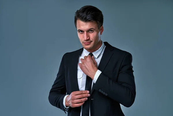 Close-up handsome and successful man in an expensive suit. He is in a white shirt with a tie. The man straightens his tie against gray background.