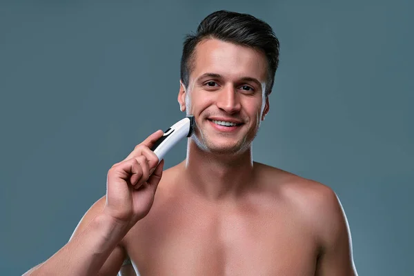 Handsome young man isolated. Portrait of shirtless muscular man is standing on gray background with trimmer in hand while shaving. Man care concept.