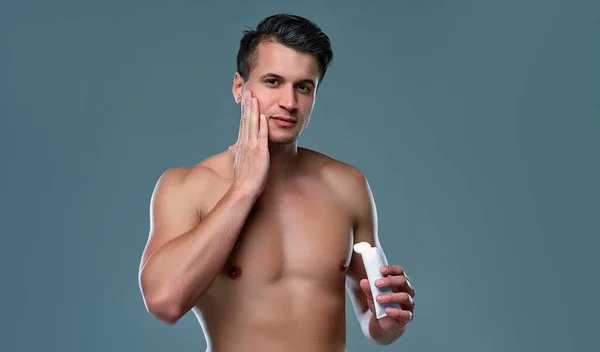 Handsome young man isolated. Portrait of shirtless muscular man is standing on grey background and using face cream. Men care concept.