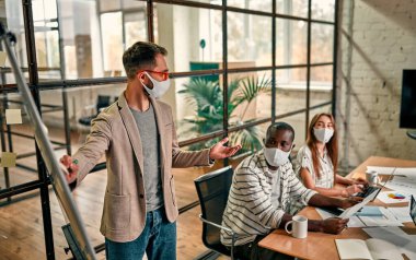 Workers discuss and meet while wearing a medical mask to protect against the corona virus. A young man writes a mind map on a whiteboard and makes new business plans during the COVID-19 pandemic.