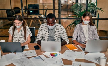 Ofis işleri bir salgın ve karantinada. Koruyucu maskeli çok ırklı iş adamlarından oluşan genç bir grup modern bir ofiste dizüstü bilgisayarlar üzerinde çalışıyor..