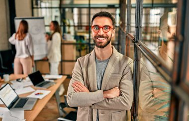 Modern bir ofiste kolları çapraz duran gözlüklü neşeli bir iş adamı. Birlikte çalışan başarılı bir hipster takımı. Bağımsızlar.