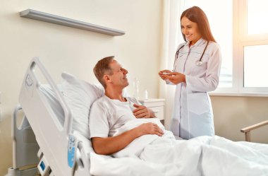 Güzel bir kadın doktor, modern bir hastane koğuşunda rehabilitasyon gören olgun bir erkek hastanın sıcaklığını ölçer..