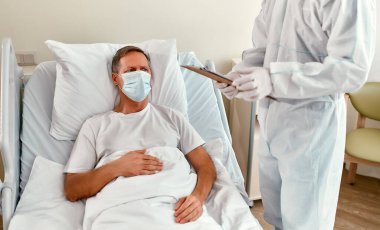 Koruyucu giysili, solunum cihazı ve gözlüklü bir doktor Coronavirüs veya covid-19 salgını sırasında modern bir hastane koğuşunda yatan olgun bir hastayı muayene eder..