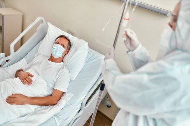 Koruyucu giysi, solunum cihazı ve gözlük takan bir doktor, Coronavirüs veya covid-19 salgını sırasında modern bir hastane koğuşunda yatan olgun bir hastaya serum damlatıyor..