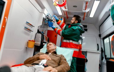 Üniformalı bir sağlık görevlisi, modern bir ambulansta sedyede yatan yaşlı bir hastaya yardım etmek için oksijen maskesi arıyor..
