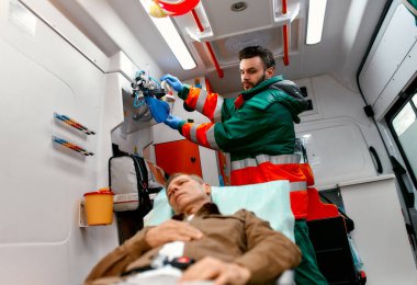 Üniformalı bir sağlık görevlisi, modern bir ambulansta sedyede yatan yaşlı bir hastaya yardım etmek için oksijen maskesi arıyor..