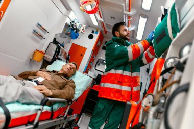 Üniformalı bir sağlık görevlisi, modern bir ambulansta sedyede yatan yaşlı bir hastaya yardım etmek için oksijen maskesi arıyor..