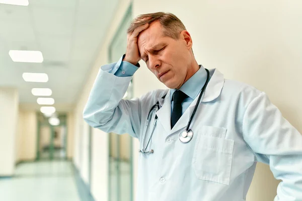 An elderly handsome upset doctor in a medical uniform with a stethoscope stands in the corridor of the clinic and put his hand on his head as a sign of despair.