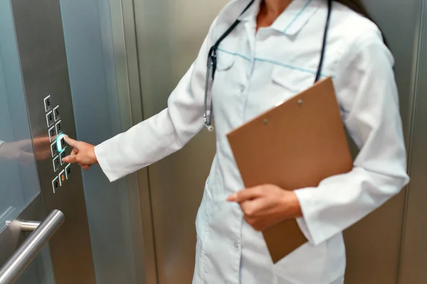 Young beautiful caucasian doctor or nurse with a stethoscope in a medical uniform and a patient card in a modern elevator presses the floor button.