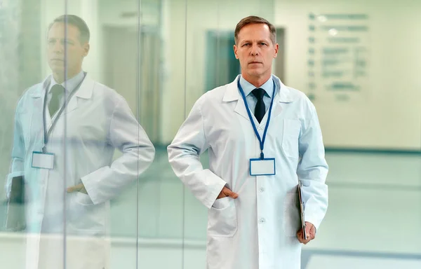 An elderly, serious, self-confident doctor in a white coat and tie with a badge stands in the corridor of the clinic.