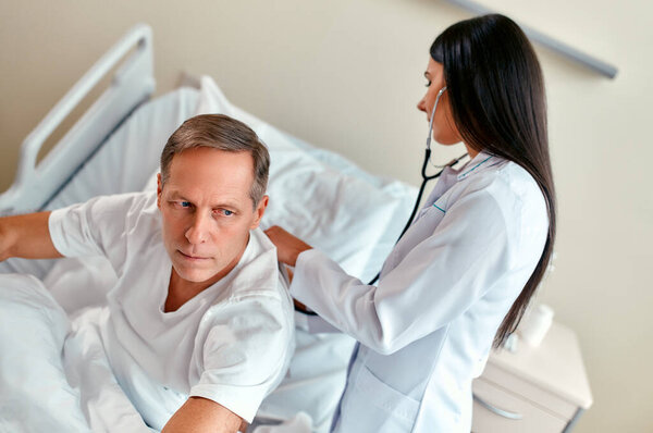 A beautiful young nurse checks the lungs of a mature patient with a stethoscope, lies on a bed in a modern ward.