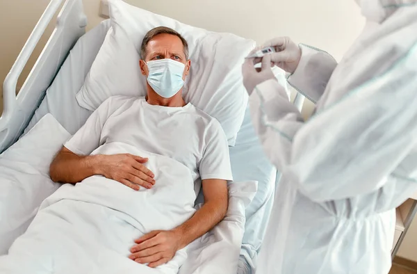 一名身穿防护服 头戴呼吸器和护目镜的医生对一名在大肠病毒或眼镜蛇流行病流行期间躺在现代医院病房里的成年病人进行检查 — 图库照片