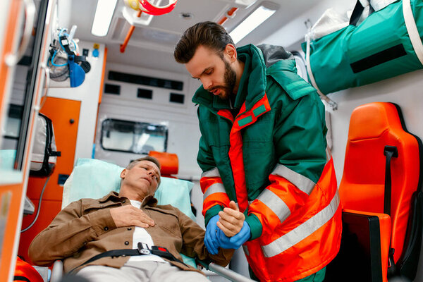 A male paramedic in uniform measures the pulse of a senior patient lying on a gurney in a modern ambulance.