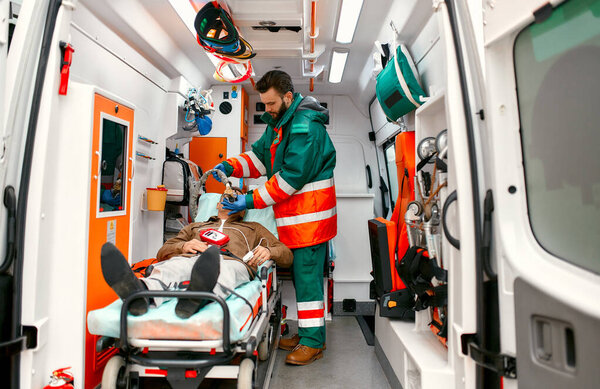 A male paramedic in uniform puts on a ventilator with oxygen to help a senior patient lying on a gurney with a pulse oximeter in a modern ambulance.