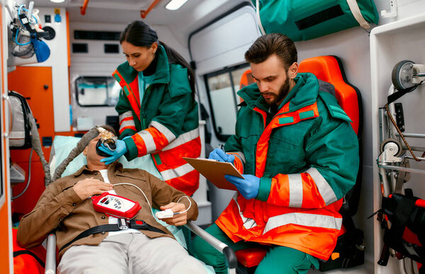 A paramedic woman in uniform puts on a ventilator with oxygen to help a senior patient lying with a pulse oximeter on a gurney in a modern ambulance. A male paramedic looks at a patient's chart.