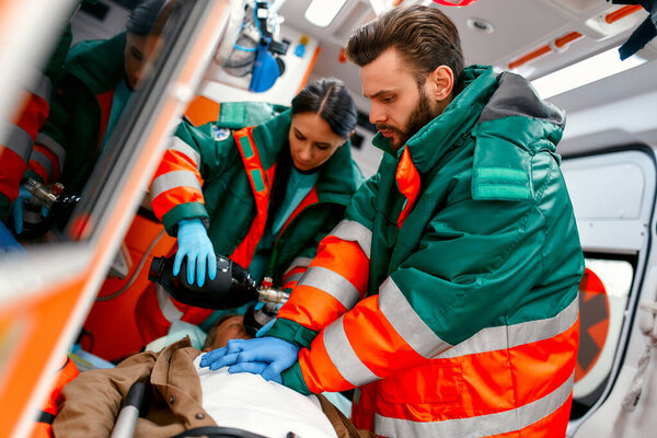 Two paramedics are resuscitating a senior lying on a gurney in an ambulance by performing chest compressions and connecting to a ventilator.