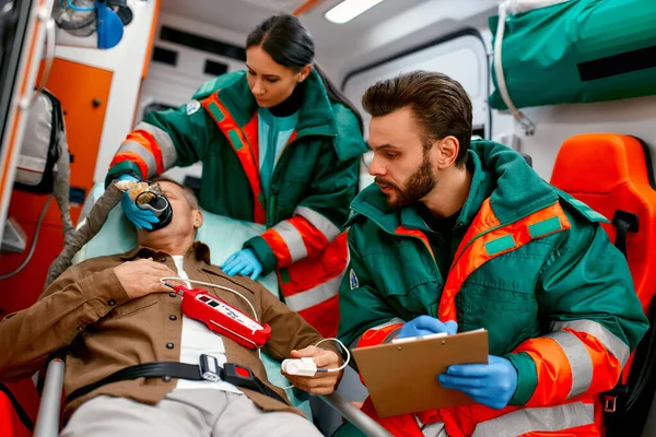 A paramedic woman in uniform puts on a ventilator with oxygen to help senior patient lying with a pulse oximeter on a gurney in modern ambulance. The male paramedic takes notes on the patient\'s card.