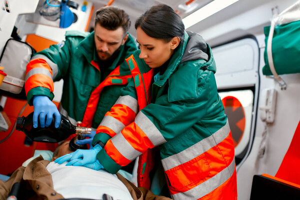 Two paramedics are resuscitating a senior lying on a gurney in an ambulance by performing chest compressions and connecting to a ventilator.