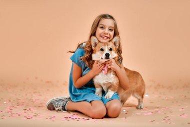 Kıvırcık saçlı tatlı bir kız yerde konfetiyle oturuyor ve boynunda fiyonk olan bir köpeğe sarılıyor..