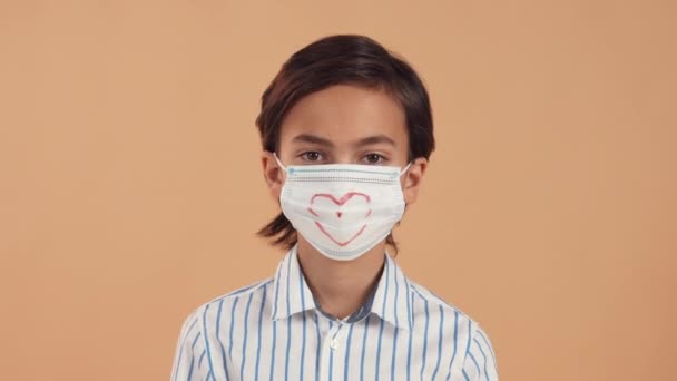 Um menino caucasiano bonito em uma camisa listrada branca e em uma máscara protetora médica com um coração desenhado mostra um gesto cardíaco com as mãos isoladas em um fundo suave pêssego. — Vídeo de Stock