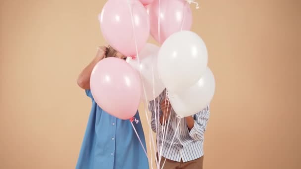 Een schattige jongen in een gestreept shirt en een schattig meisje met krullend haar in een blauwe jurk gluur uit van achter ballonnen met valentijnskaarten harten in handen geïsoleerd op een perzik zachte achtergrond. — Stockvideo