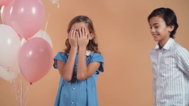 En söt pojke i randig skjorta ger en låda med en gåva till en söt flicka med lockigt hår i en blå klänning isolerad på en persika delikat bakgrund. — Stockvideo